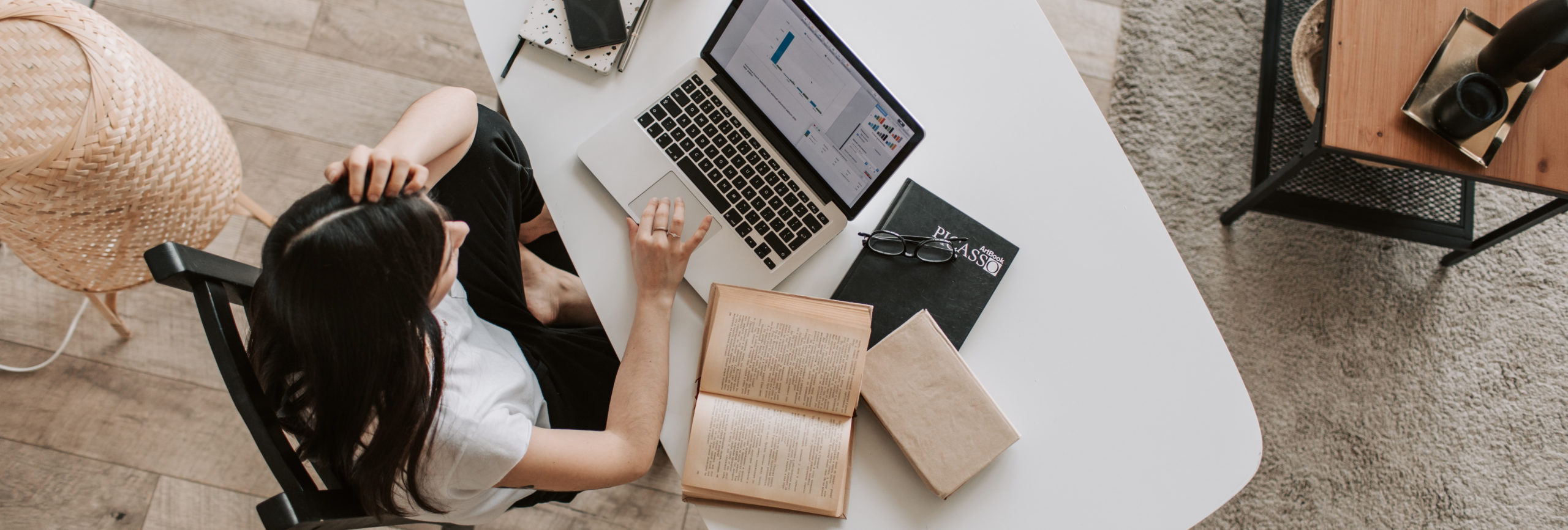 top view of a female looking at her laptop with an open book slightly to the side of the laptop