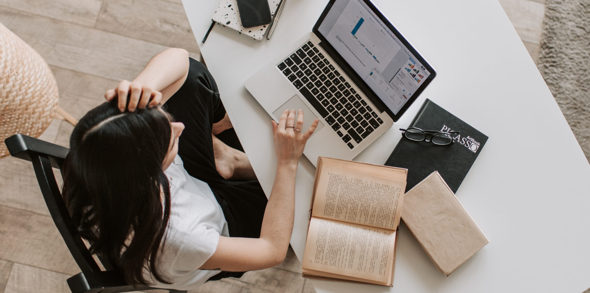 top view of a female looking at her laptop with an open book slightly to the side of the laptop